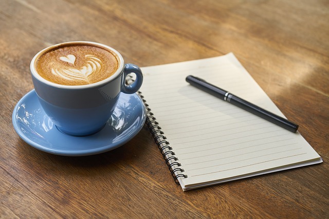 Un carnet, un stylo et une tasse de cappuccino, posés sur un bureau en bois.